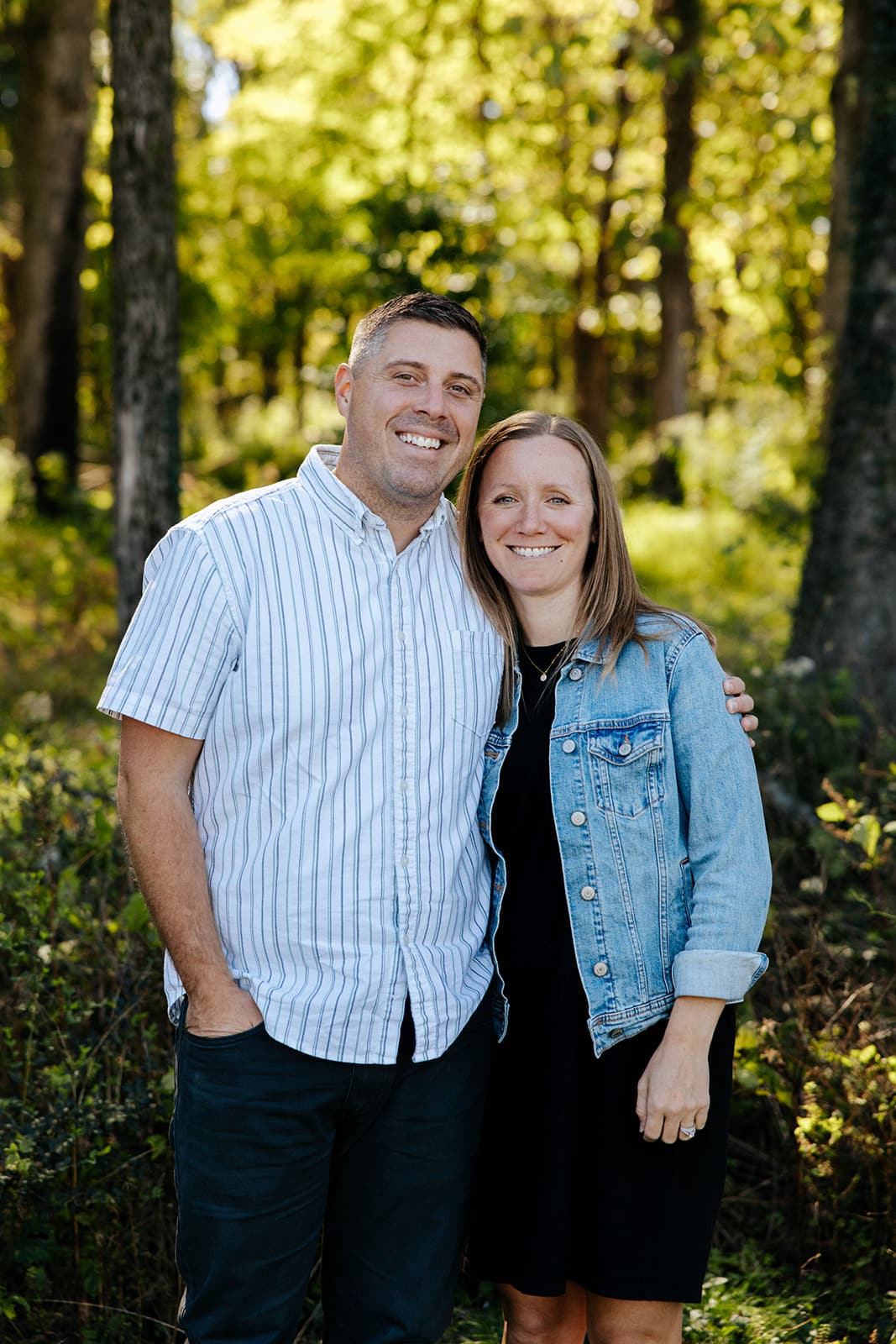 Smiling couple stands together in a sunlit forest, man in a striped shirt and woman in a denim jacket with trees in the background.
